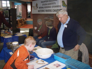 Clyde Marsteller standing across table from boy who is looking at drawings and samples of invertebrates.