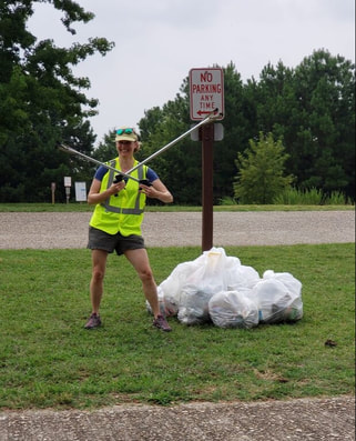 volunteer standing by a road, next to filled trash bags and holding two litter grabbers 