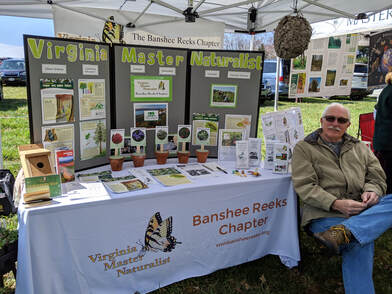 Man sits at a booth at a festival with a display about plants, birds, and trees.