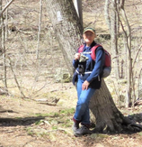 person with binoculars and backpack leaning against a tree