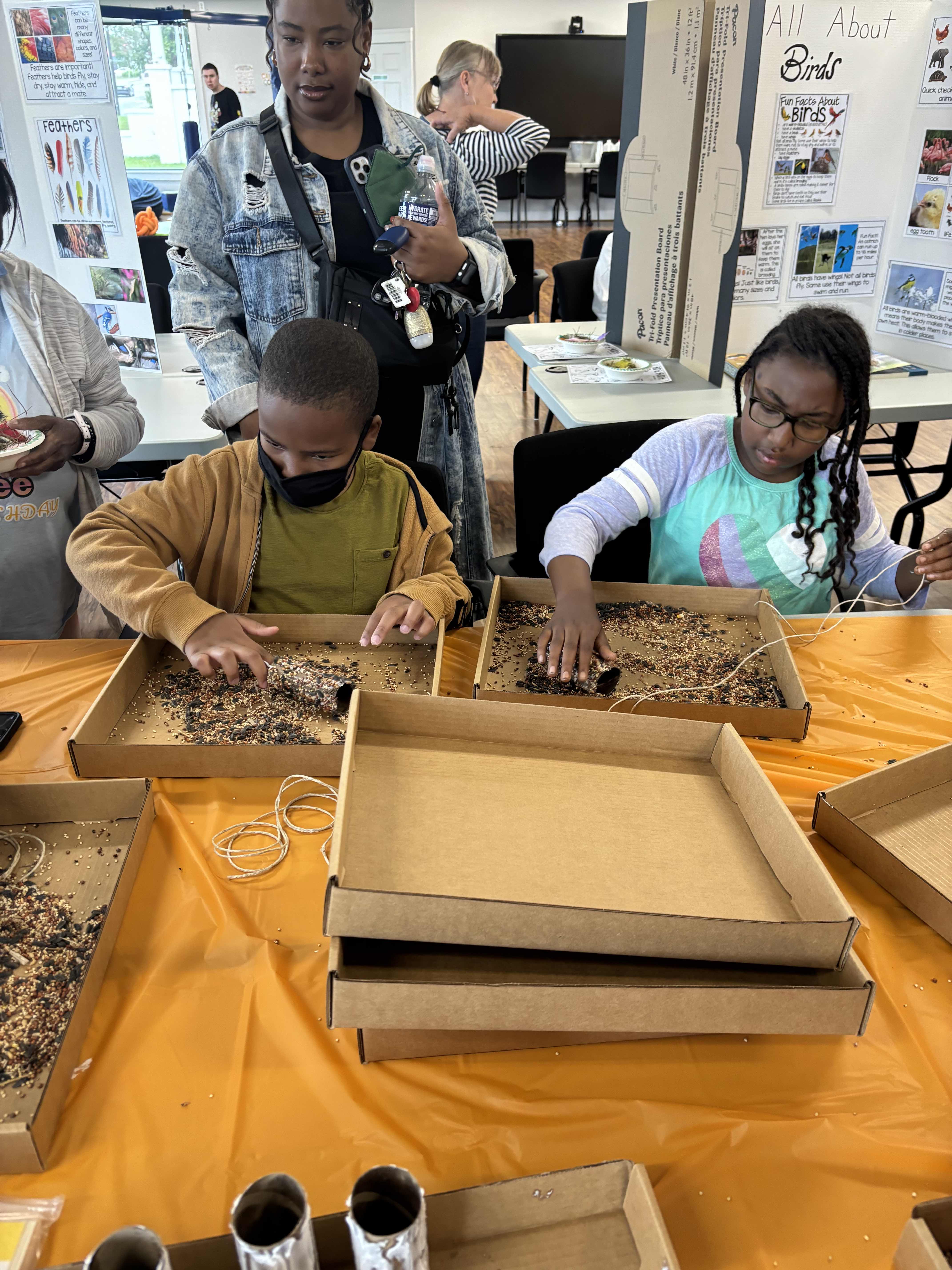 two children touching soil in cardboard trays as an adult looks over their shoulders
