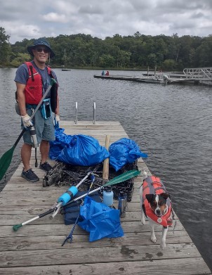 volunteer posed on a dock with bags of trash and a dog wearing a life preserver