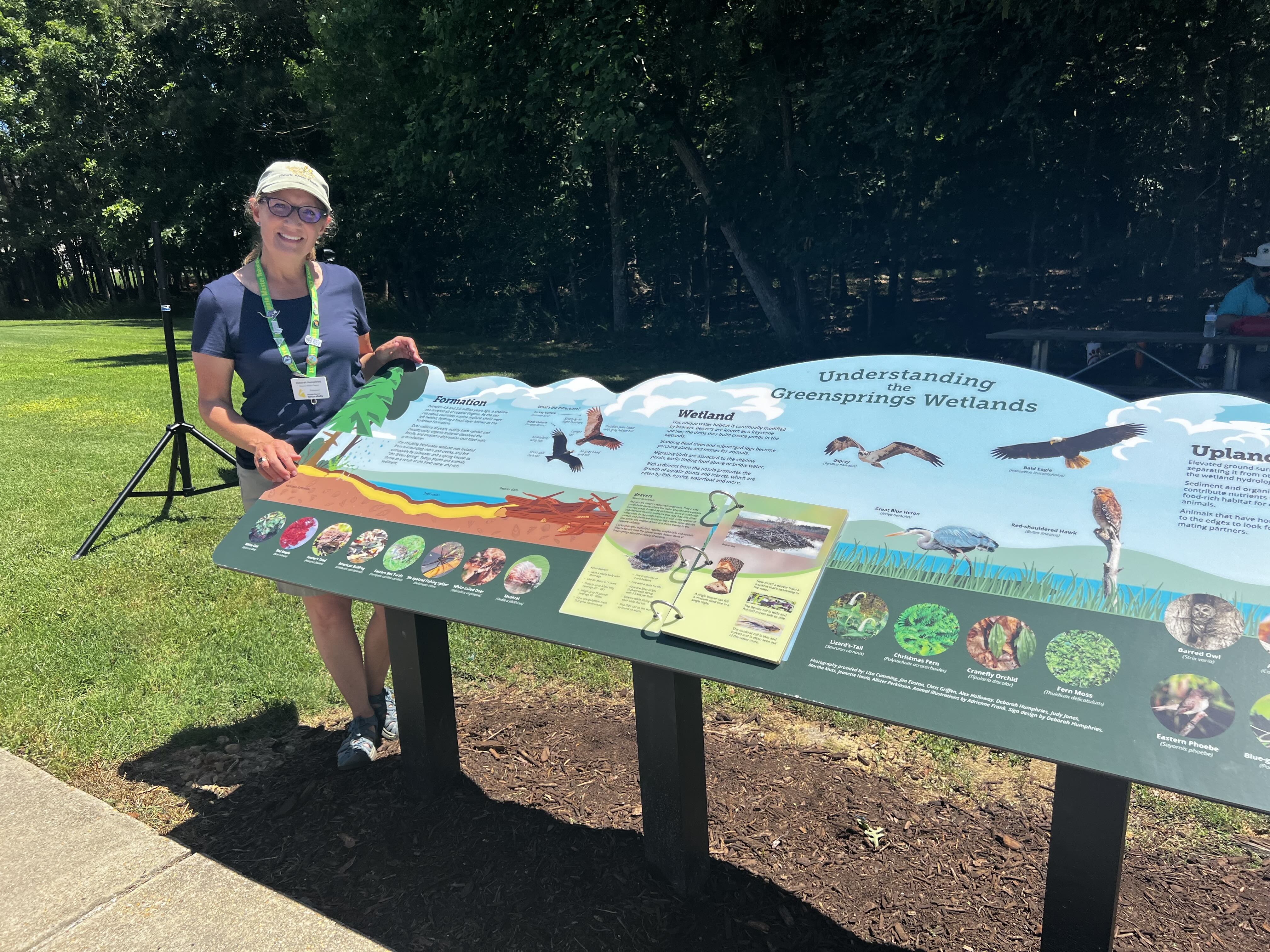 volunteer posed with a colorful interpretive sign about understanding the Greenspring wetlands
