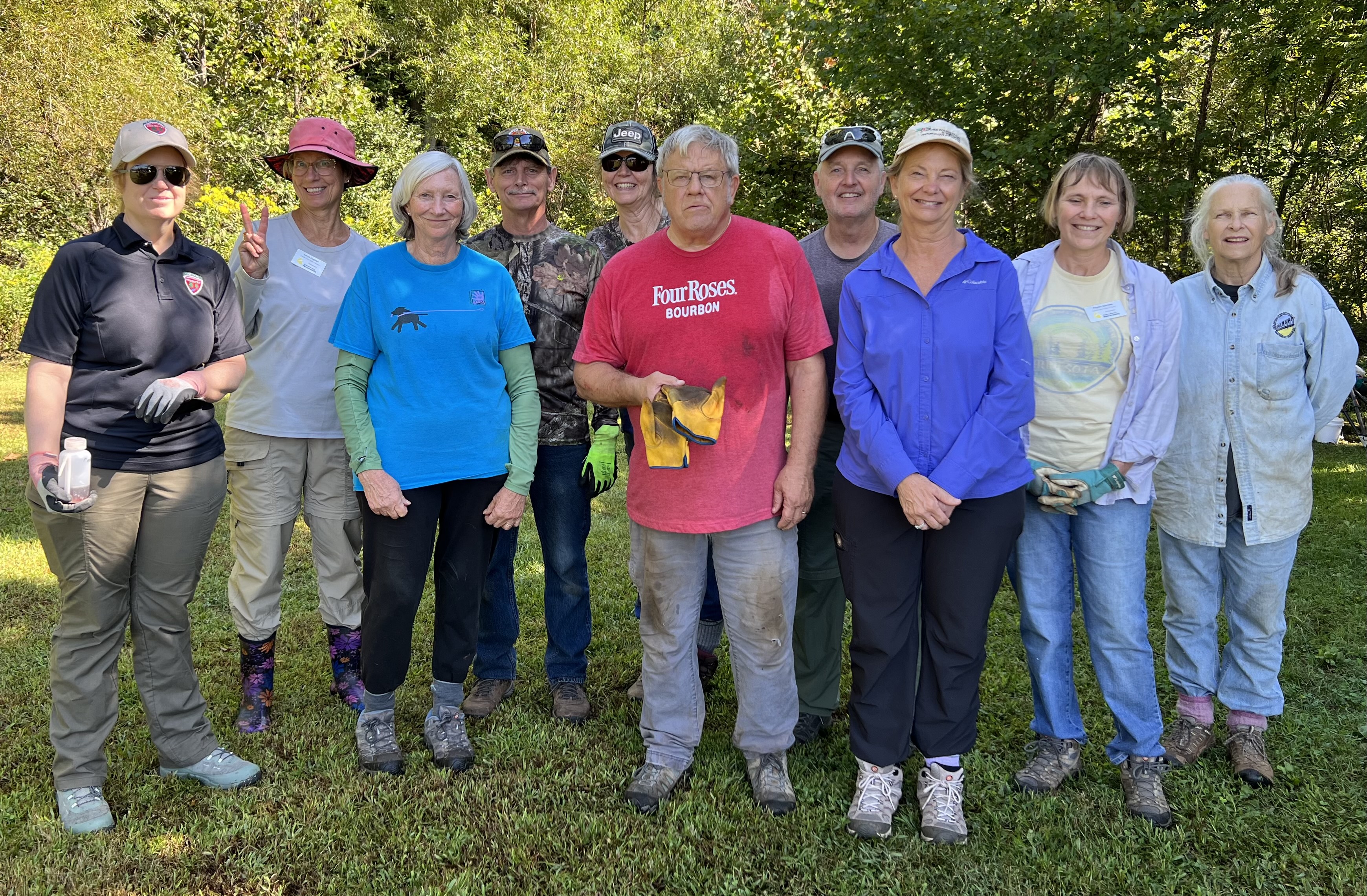 ten people wearing gardening clothes and some in work gloves, posed outdoors in from of trees on a sunny day