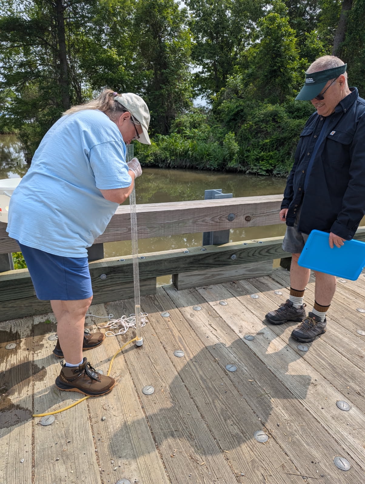 two people on a wooden dock near water. One person is holding a clipboard. The other person is peering into a tall clear tube.