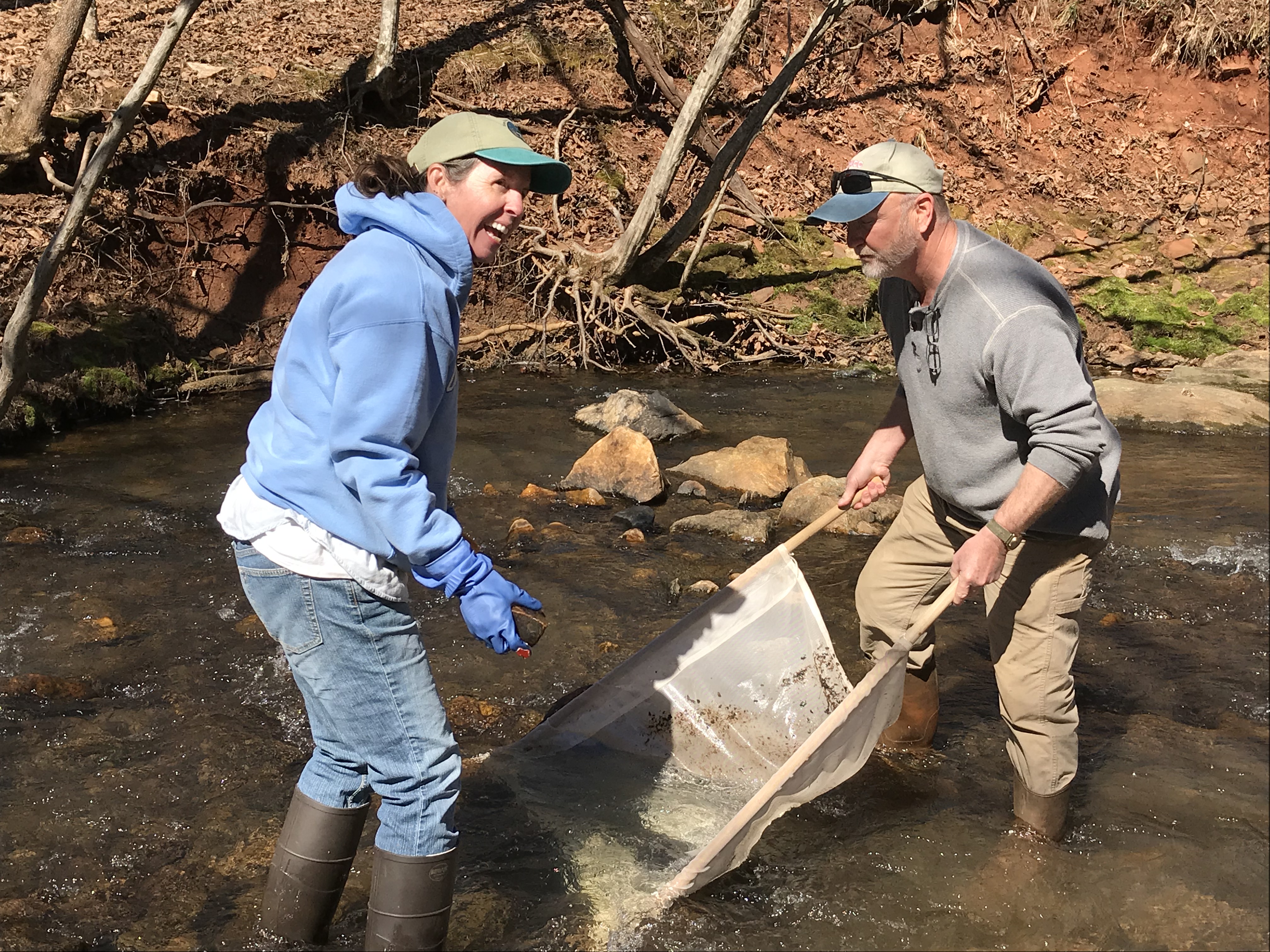 two people smiling, wearing waders and using a kick net in a rocky stream on a sunny day