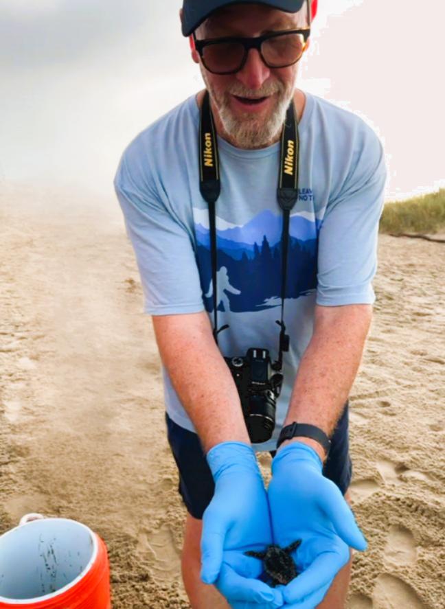 A man wearing a ball cap, with a camera around his neck, holds his hands out and overlapped in front of him. In his palms is a teeny tiny baby turtle.