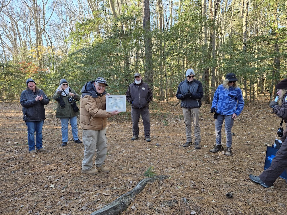 man in winter jacket holds a certificate of appreciation while surrounded by clapping people in a forest clearing