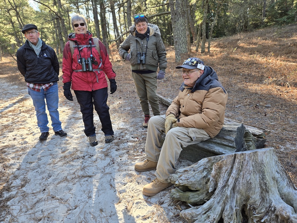 man in winter coat seated on a log in a sandy wooded area, with three other people posed nearby