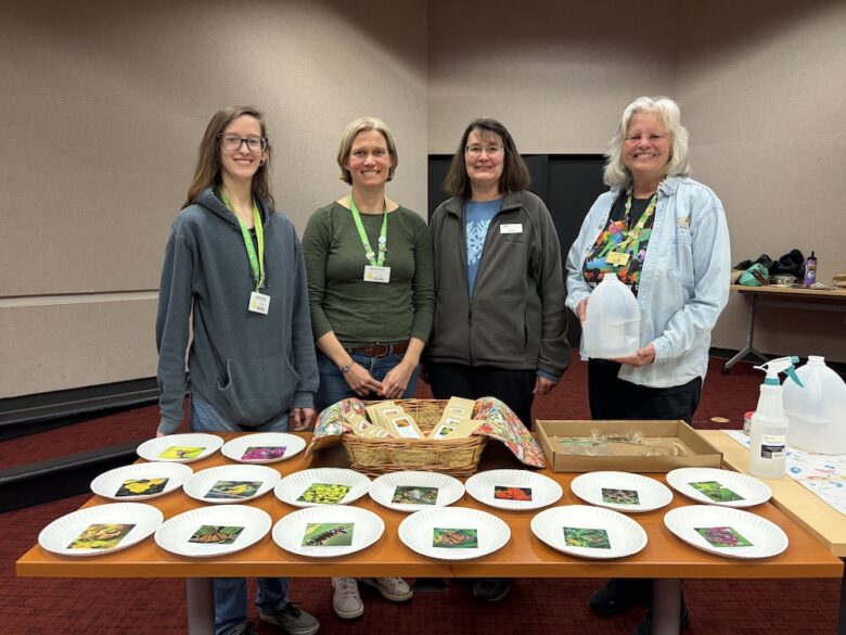 four volunteers pose in front of a table with paper plates that have pictures of butterflies and caterpillars on them