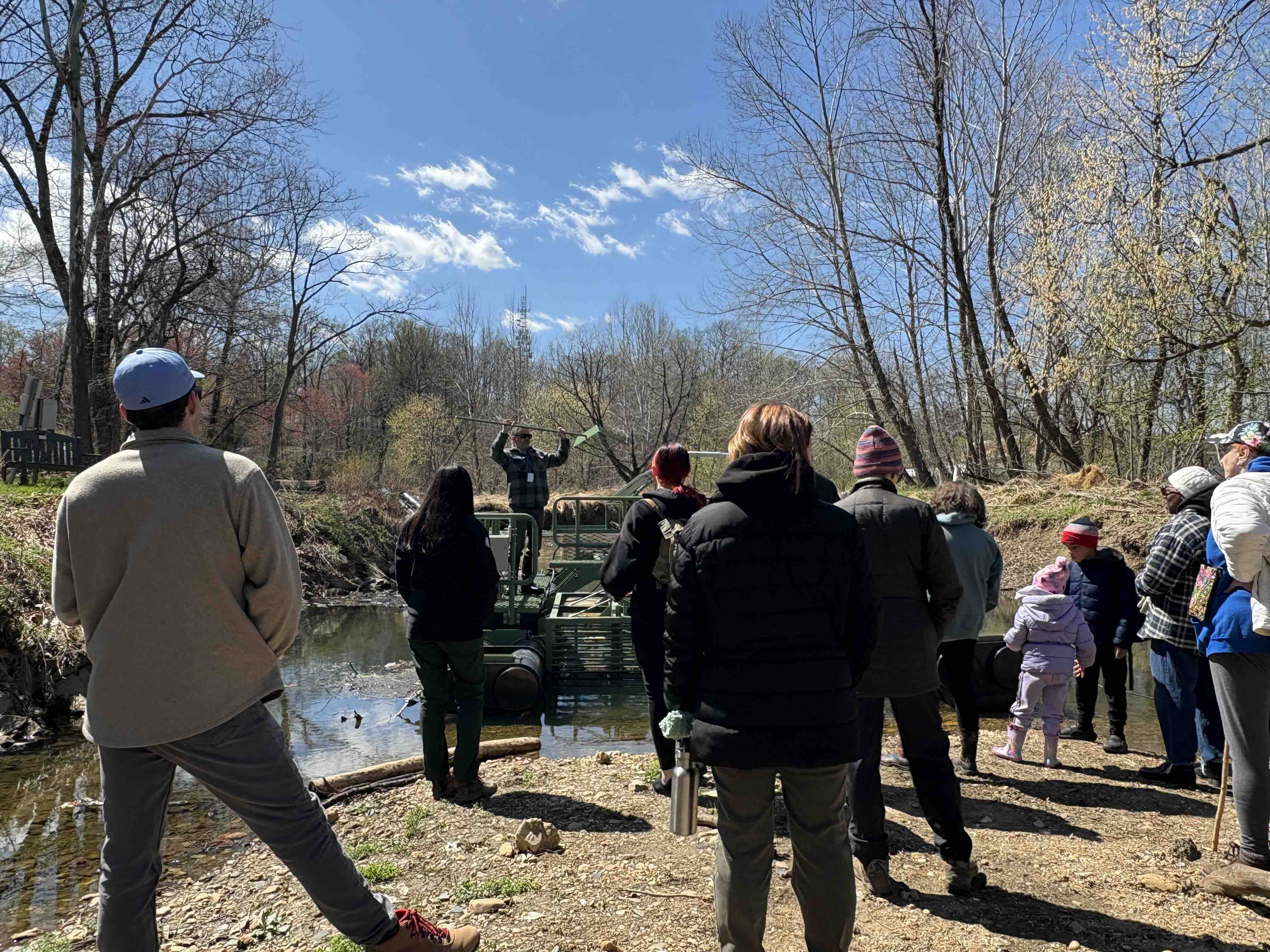 group of people outdoors watching a demonstration of a large machine that removes trash from a stream