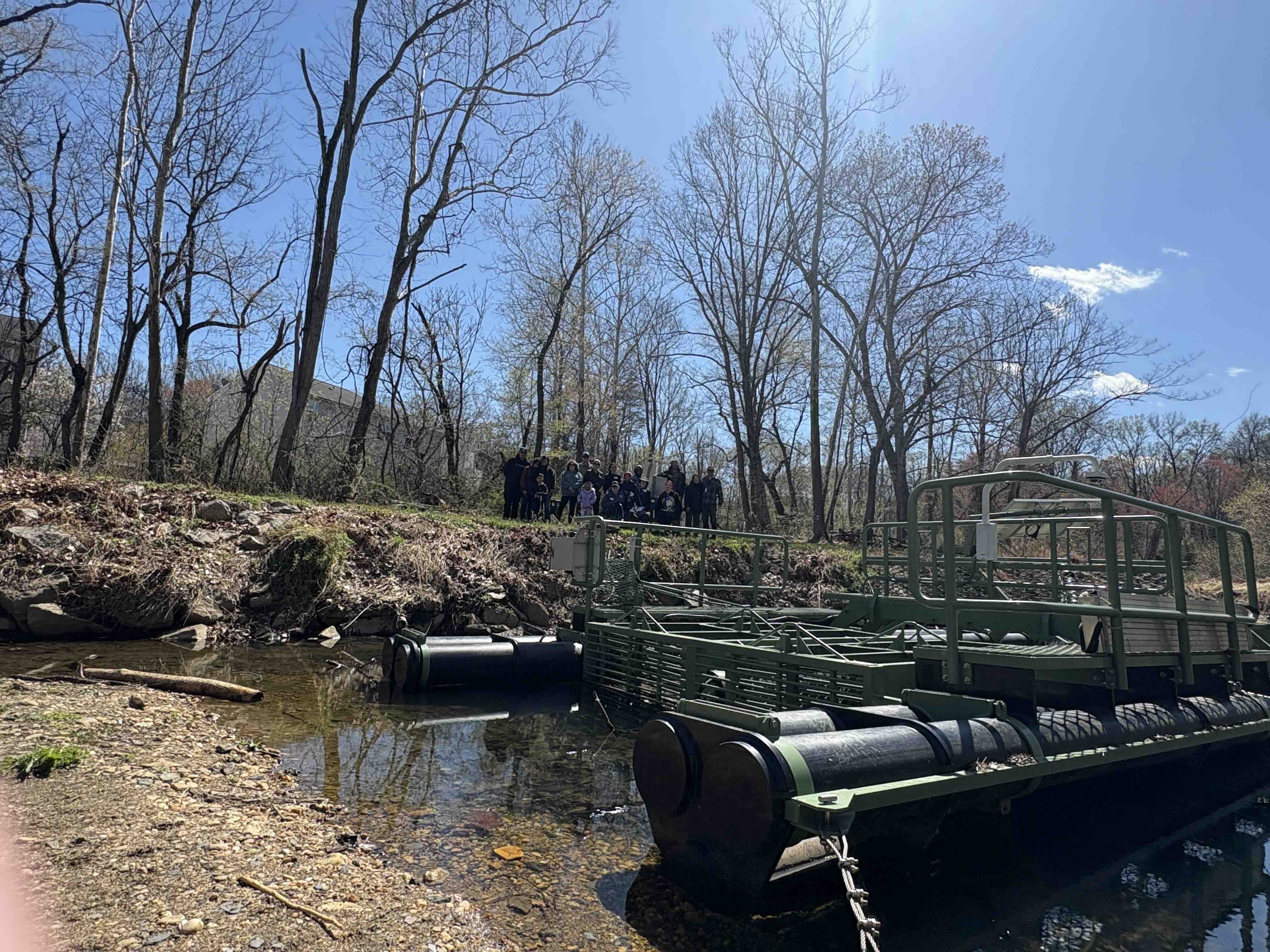 large machine in a stream with a group of people posed behind it in the distance