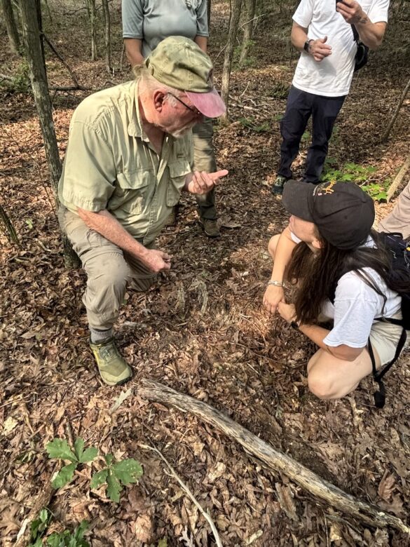 volunteer shows a mushroom to a person as they both kneel in a forest
