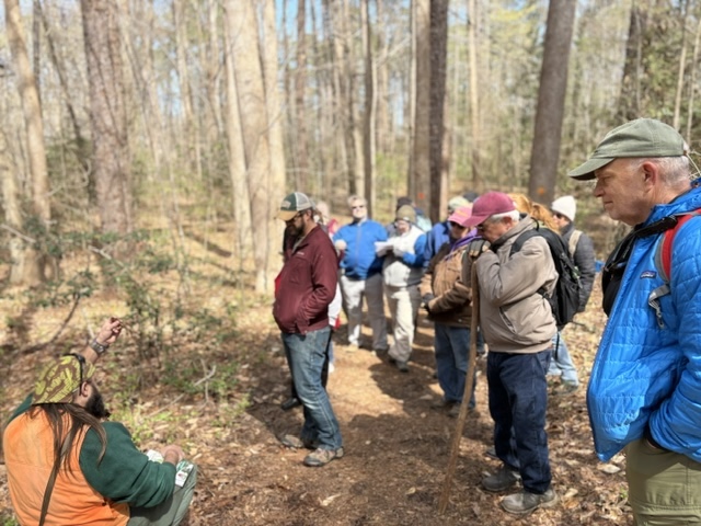 people line up along a forest trail, listening to an instructor who is kneeling on the ground