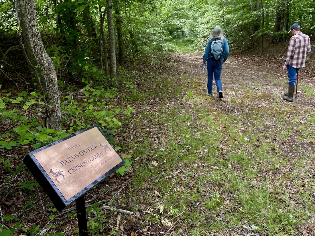 Mounted plaque in a forest reading "Here is Patawomeck Cepsiin (Land)". Two people are walking down the trail near the plaque.